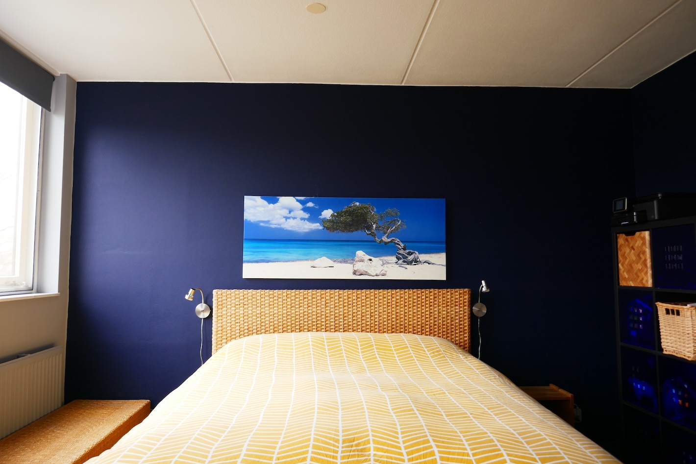 Een slaapkamer met een donkerblauwe muur en een bed met een geweven hoofdbord. Boven het bed hangt een schilderij van een strand met een eenzame boom. Het bed heeft een geel en wit dekbedovertrek.