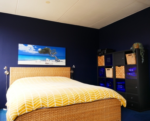 Dezelfde slaapkamer vanuit een andere hoek, met een donkerblauwe muur en een bed met een geweven hoofdbord. Er staat een zwarte opbergkast met rieten manden aan de rechterkant van de kamer.