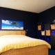 Dezelfde slaapkamer vanuit een andere hoek, met een donkerblauwe muur en een bed met een geweven hoofdbord. Er staat een zwarte opbergkast met rieten manden aan de rechterkant van de kamer.